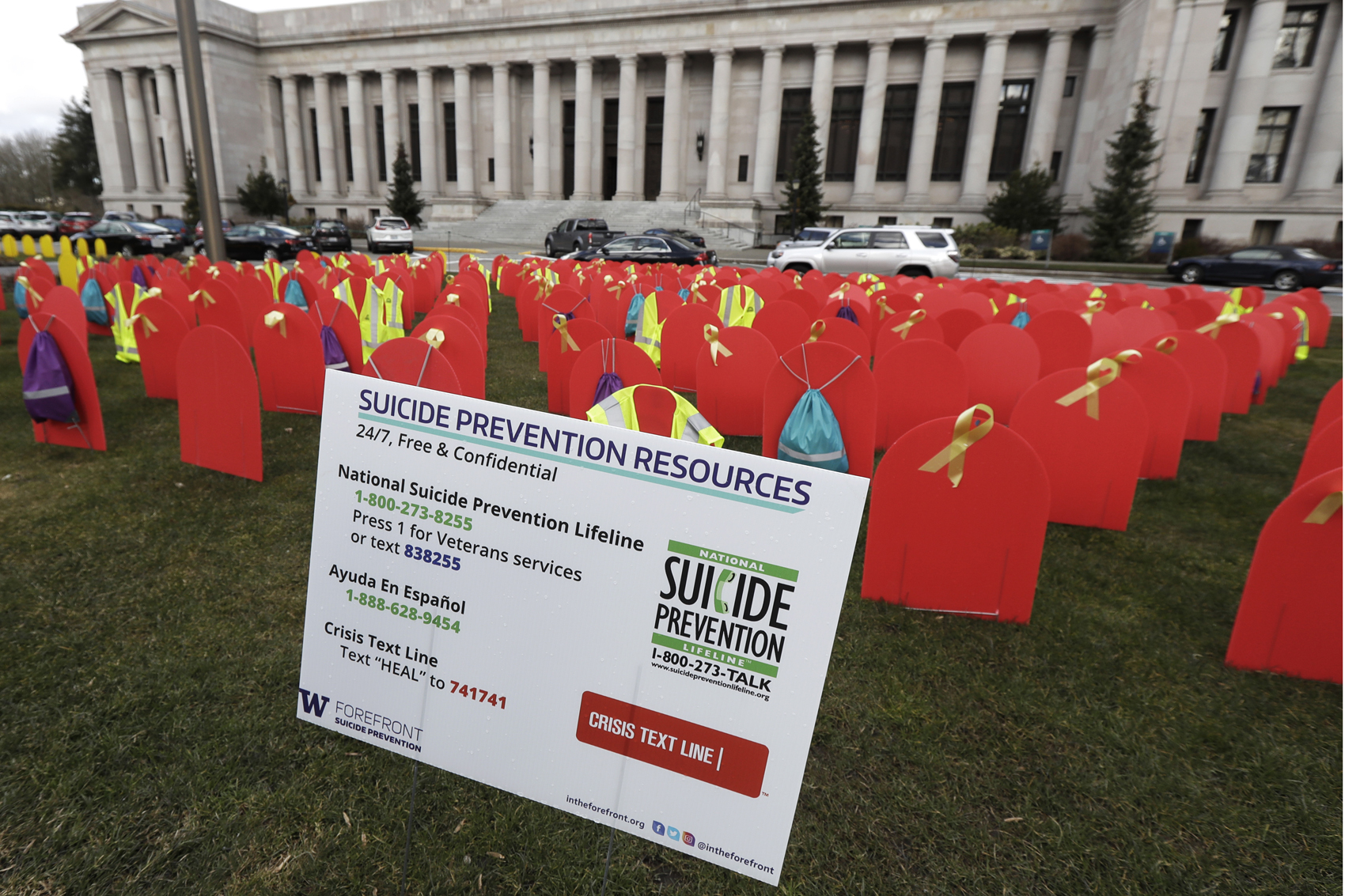 caption: Suicide prevention phone numbers and red mock tombstones designating some of the more than 1,000 people who took their lives by suicide in Washington state in 2017 are displayed on a grassy area near the Temple of Justice, Tuesday, March 12, 2019, at the Capitol in Olympia, Wash. (Ted S. Warren/AP)