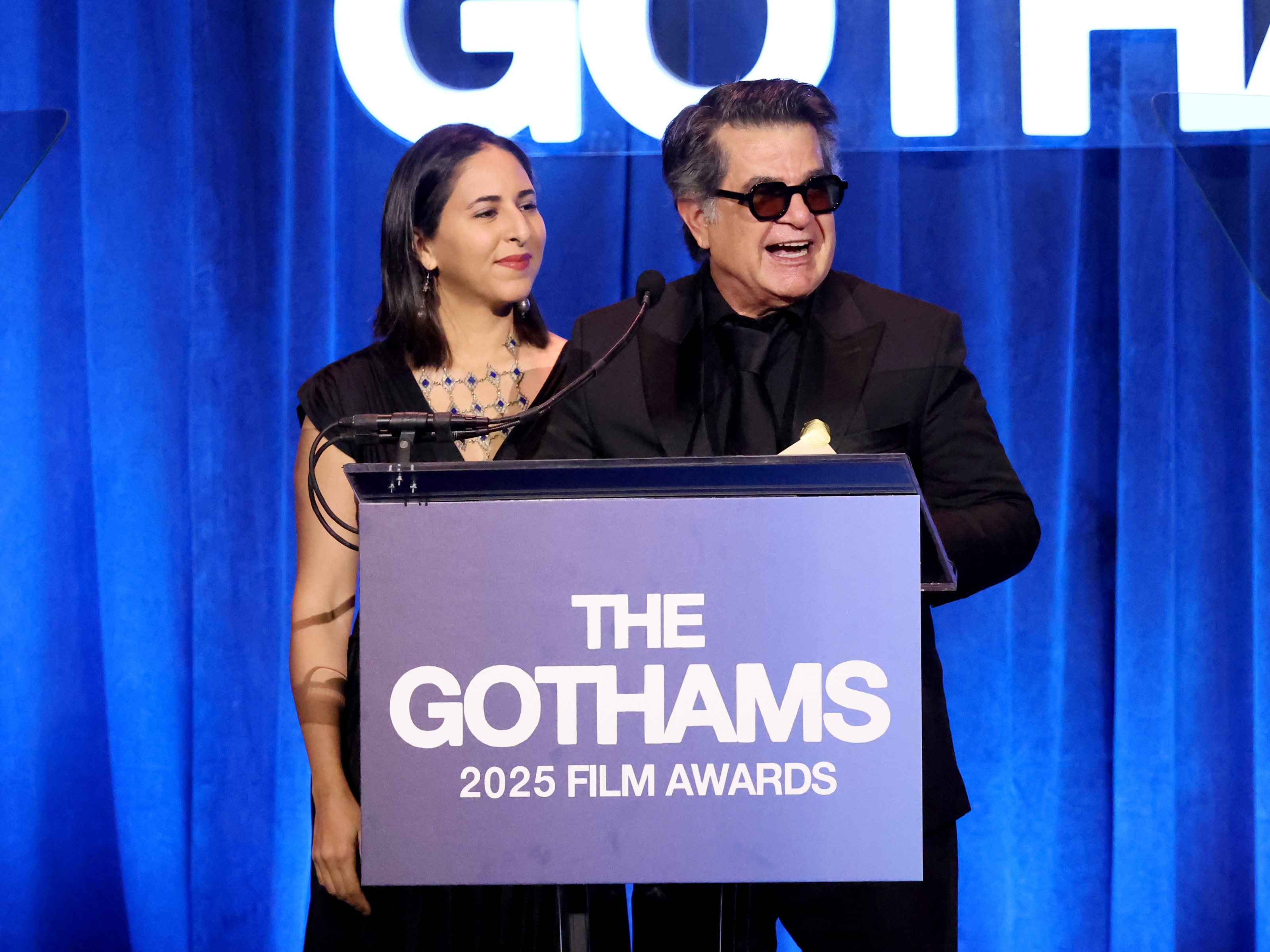 caption: Alongside interpreter Sheida Dayani, filmmaker Jafar Panahi accepts the award for the best original screenplay at the Gotham Awards in New York on Monday for his film <em>It Was Just an Accident</em>.