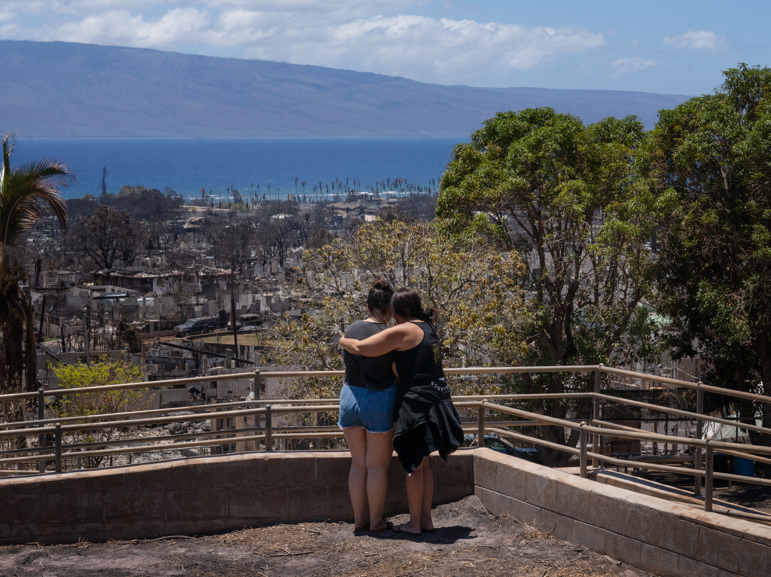 caption: Two women embrace and cry as they look out over a burned area in Lahaina, Hawaii in August 2023. A new survey finds most Americans expect the impacts of climate change to worsen in the next 30 years, as climate scientists warn.