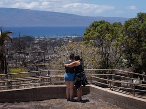 caption: Two women embrace and cry as they look out over a burned area in Lahaina, Hawaii in August 2023. A new survey finds most Americans expect the impacts of climate change to worsen in the next 30 years, as climate scientists warn.