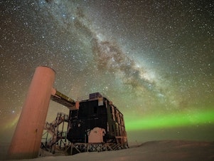 caption: The IceCube Lab under a starry, night sky, with the Milky Way appearing over low auroras in the background.
