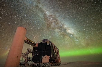 caption: The IceCube Lab under a starry, night sky, with the Milky Way appearing over low auroras in the background.