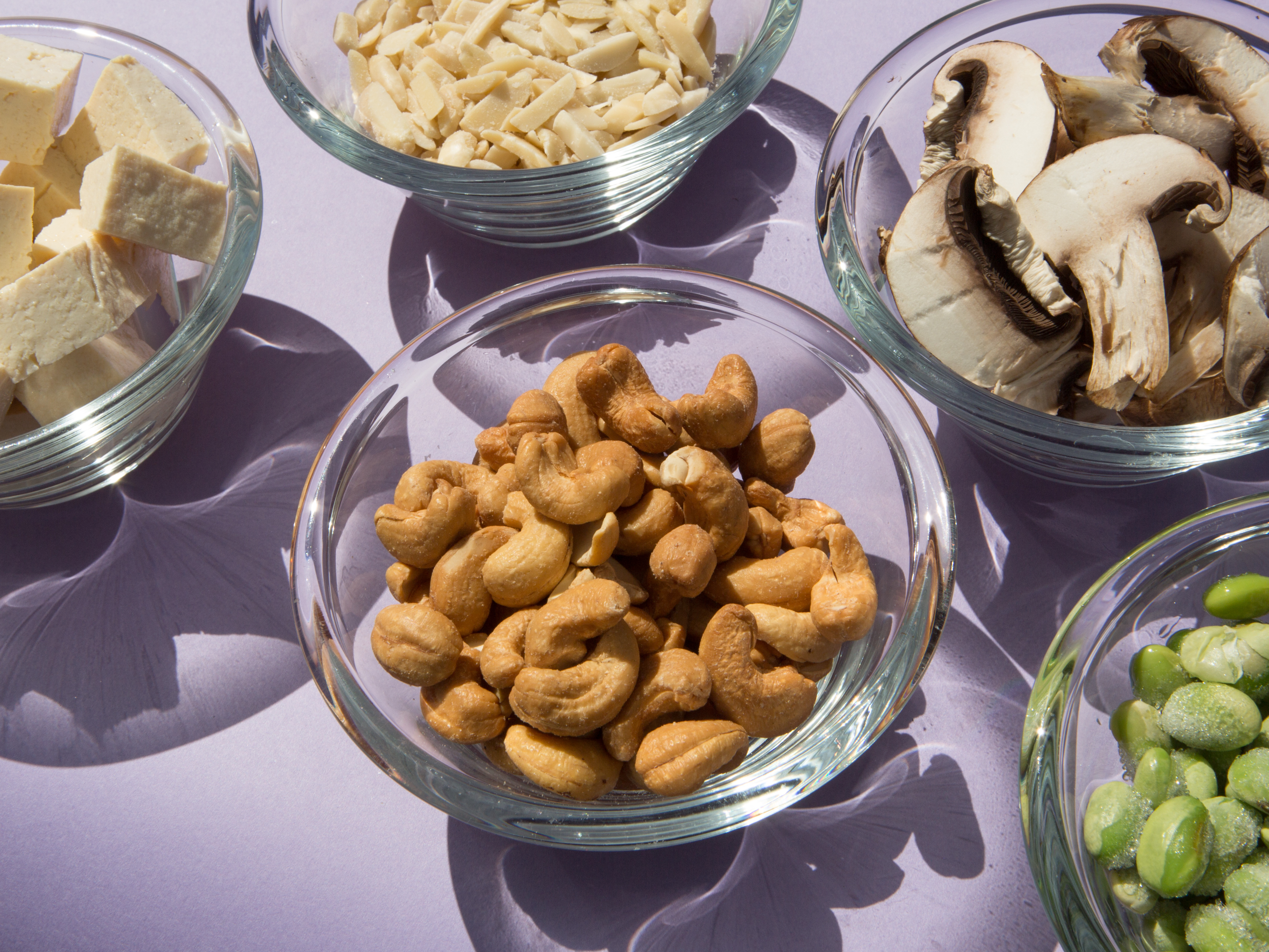 Image of small glass bowls filled with tofu, sliced almonds, cashews, sliced mushrooms and frozen edamame. The bowls are arranged in a cluster and the image is taken from a 3/4 angle against a light purple backdrop.