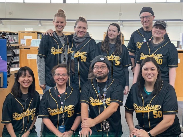 caption: The Seattle Public Library's Broadview Branch staff pose in matching baseball jerseys emblazoned with "Broadview 50" in the style of the Milwaukee Brewers' team jerseys. 