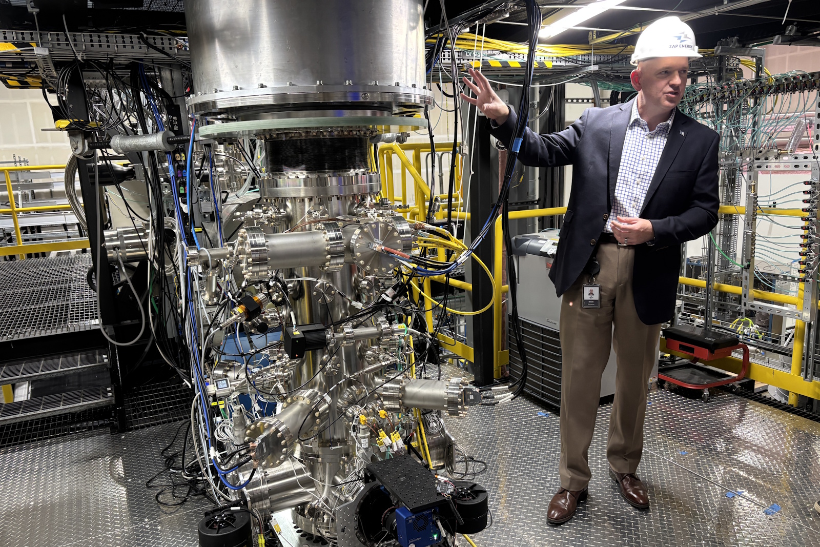 caption: Matt Thompson of Zap Energy explains how its latest reactor works, before a live demonstration. This reactor is currently firing inert gas, to monitor one stage of the fusion process, rather than fully undergoing fusion. Zap uses a patented "Z-pinch" technology, and is a few years behind Helion. Helion has been doing live fusion for several years now, and is currently working on making the process more efficient.