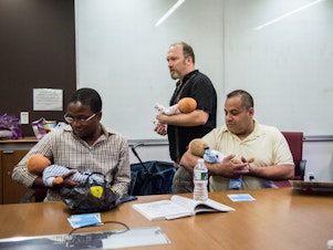 caption: Joe Bay (center), coach of a New York City "Bootcamp for New Dads," instructs Adewale Oshodi (left) and George Pasco in how to cradle an infant for best soothing.