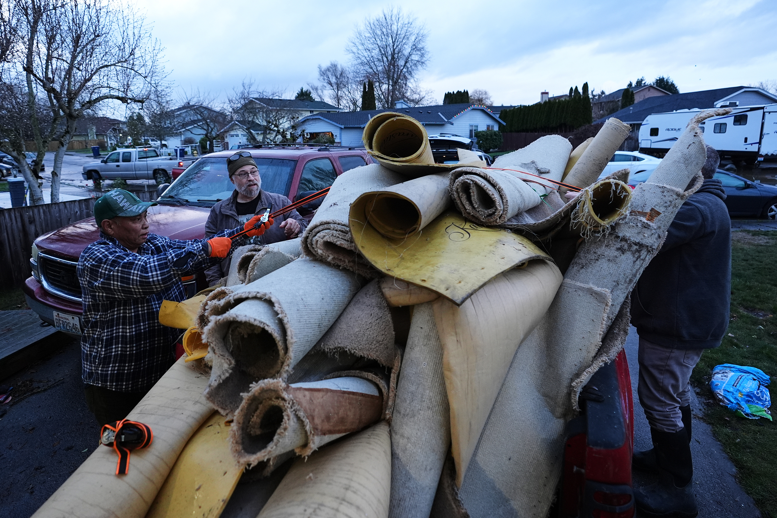 caption: Chanthy Khuon, left, and Bryan Sheriff, second from left, try to secure a large pile of carpet and padding onto a truck after taking it out of the home of Khuon's son after flooding Wednesday, Dec. 17, 2025, in Pacific, Wash. 