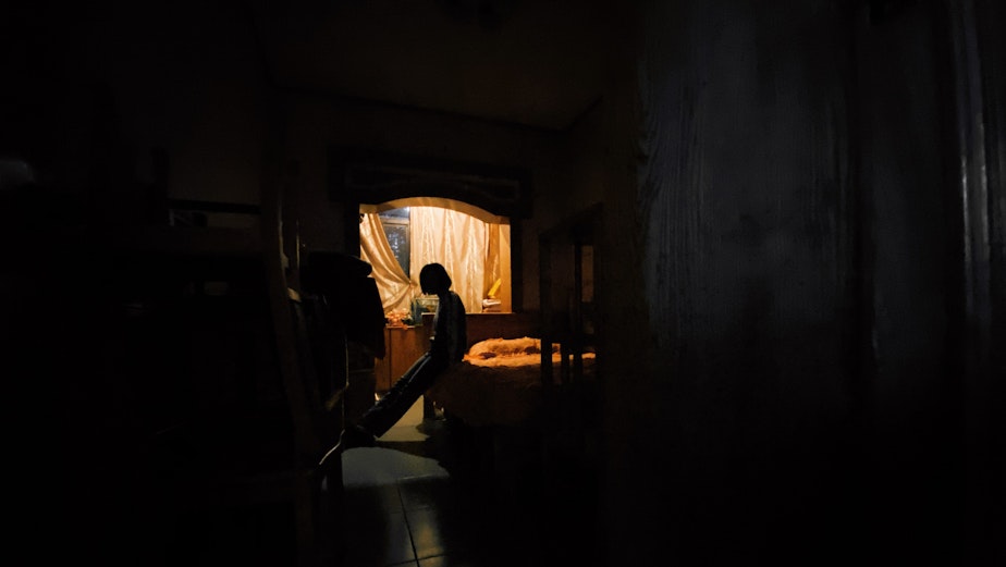 caption: An isolated person leans against their bed. They look outside a window through a small opening in the curtain.
