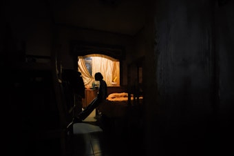 caption: An isolated person leans against their bed. They look outside a window through a small opening in the curtain.