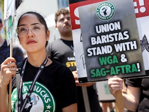 caption: Starbucks workers including barista Kat Ramos stand with striking SAG-AFTRA and Writers Guild of America members on the picket line in solidarity outside Netflix studios on July 28, 2023 in Los Angeles, Calif.