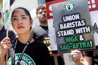 caption: Starbucks workers including barista Kat Ramos stand with striking SAG-AFTRA and Writers Guild of America members on the picket line in solidarity outside Netflix studios on July 28, 2023 in Los Angeles, Calif.