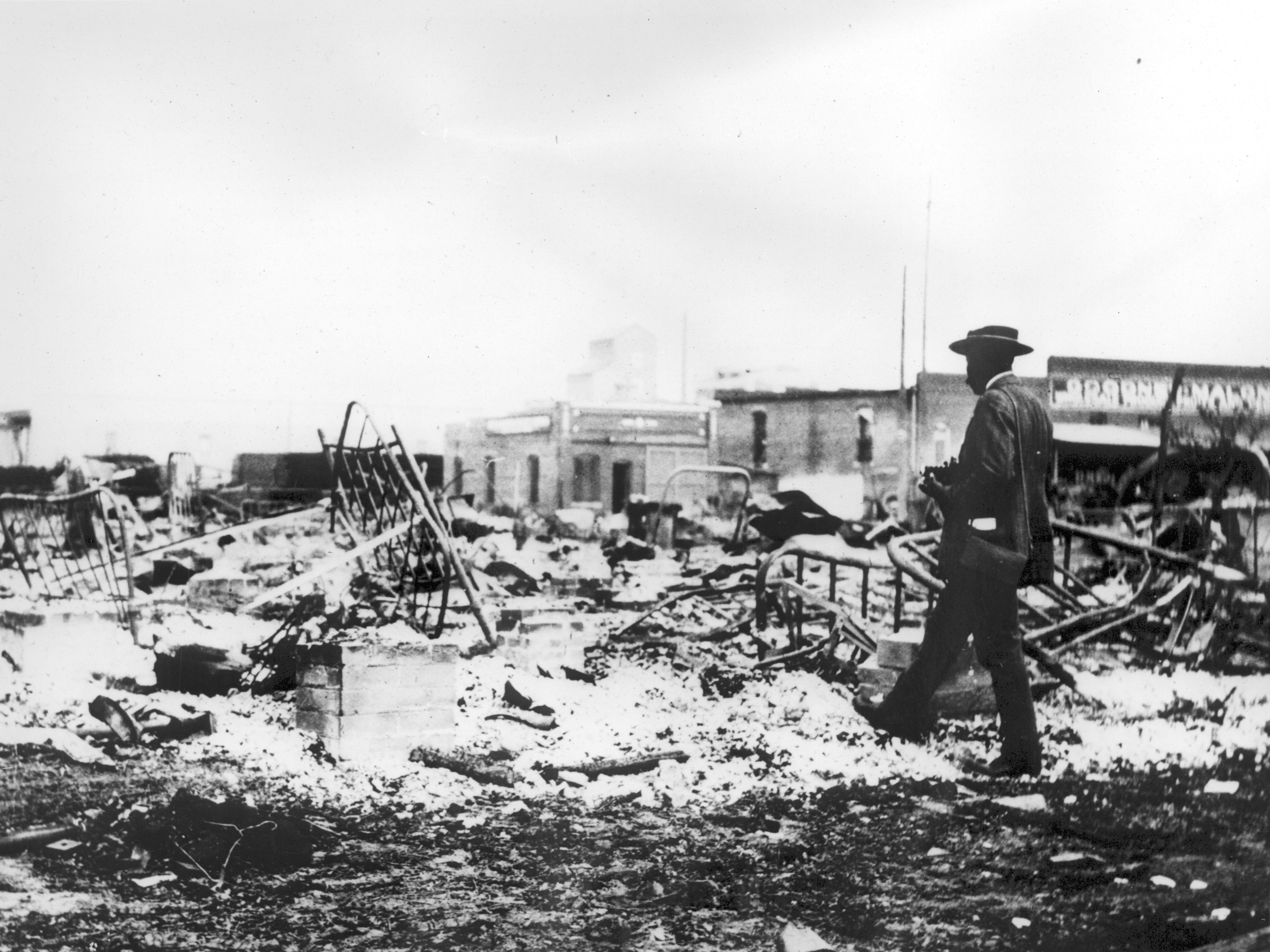 caption: Photograph of an African-American man with a camera looking at the skeletons of iron beds which rise above the ashes of a burned-out block after the 1921 Tulsa Race Massacre, in Oklahoma.