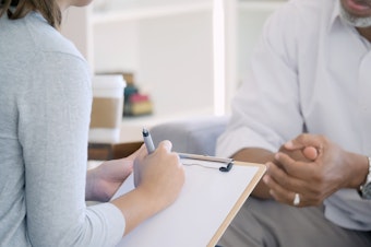 Concerned senior African American man talks with a female mental health professional. The mental health professional is taking notes in the foreground.