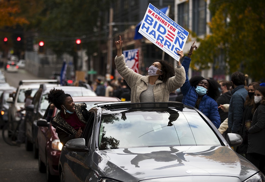caption: Oksana Reva, center, holds a Biden-Harris sign out of a sun roof while celebrating after Joe Biden was officially named the president elect on Saturday, November 7, 2020, at the intersection of 10th Avenue and East Pine Street in Seattle.