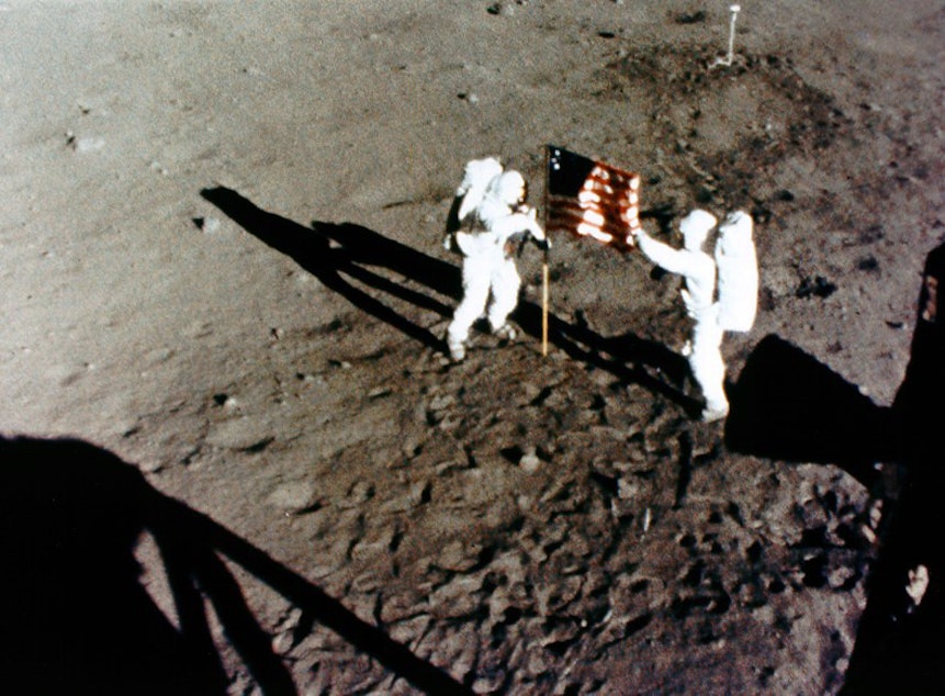 caption: This frame from the 16-mm camera mounted in the Lunar Module window shows Neil Armstrong (left) and Buzz Aldrin (right) deploying the U.S. Flag.