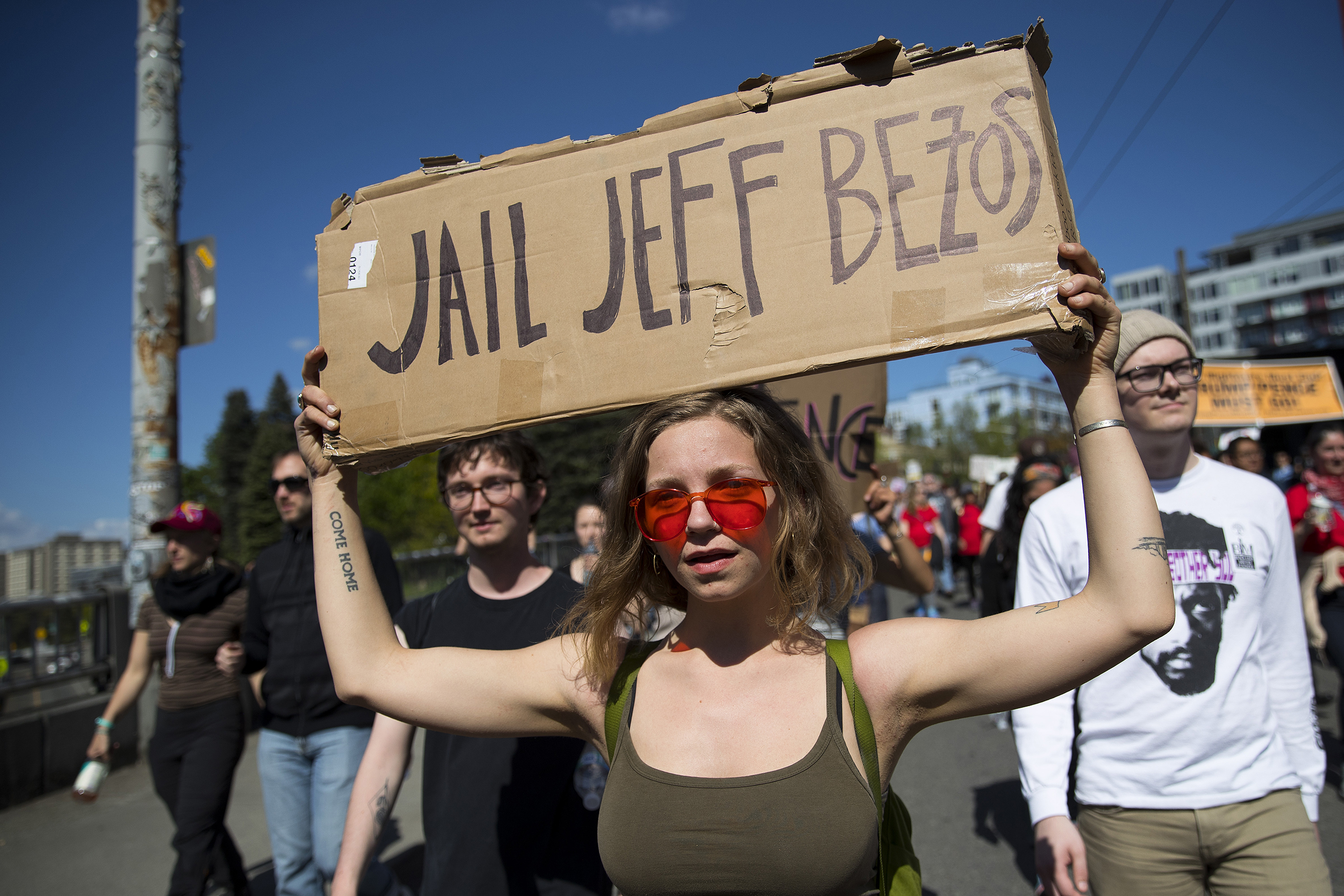 caption: Lexie Mumey, 23, carries a sign that reads 'Jail Jeff Bezos' while marching during the annual May Day march on Wednesday, May 1, 2019, in Seattle.