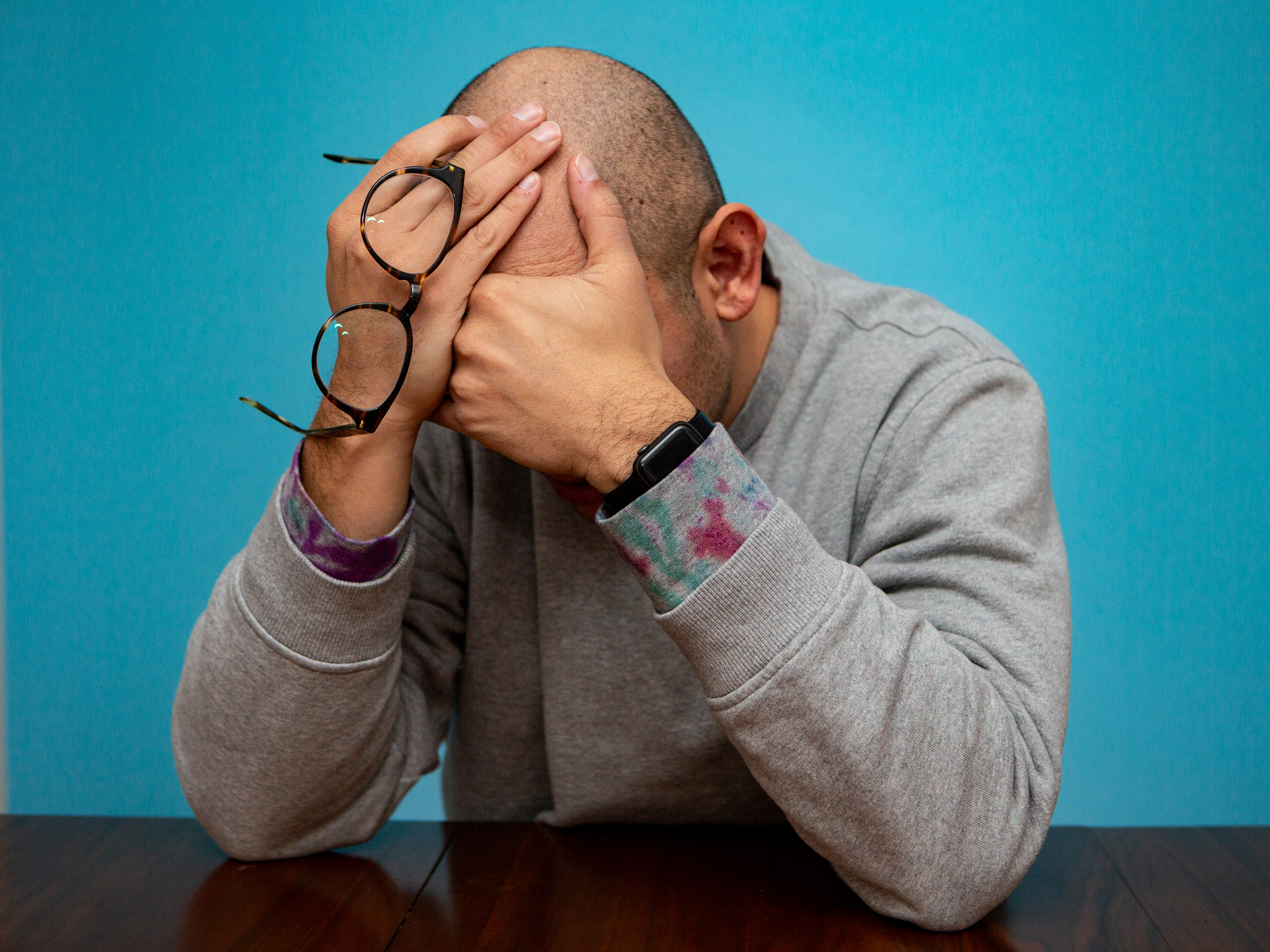 Photograph of a man sitting with his elbows on a wooden table and face palming due to distress and overwhelm. He holds his glasses in his hands. He sits in front of a blue backdrop.
