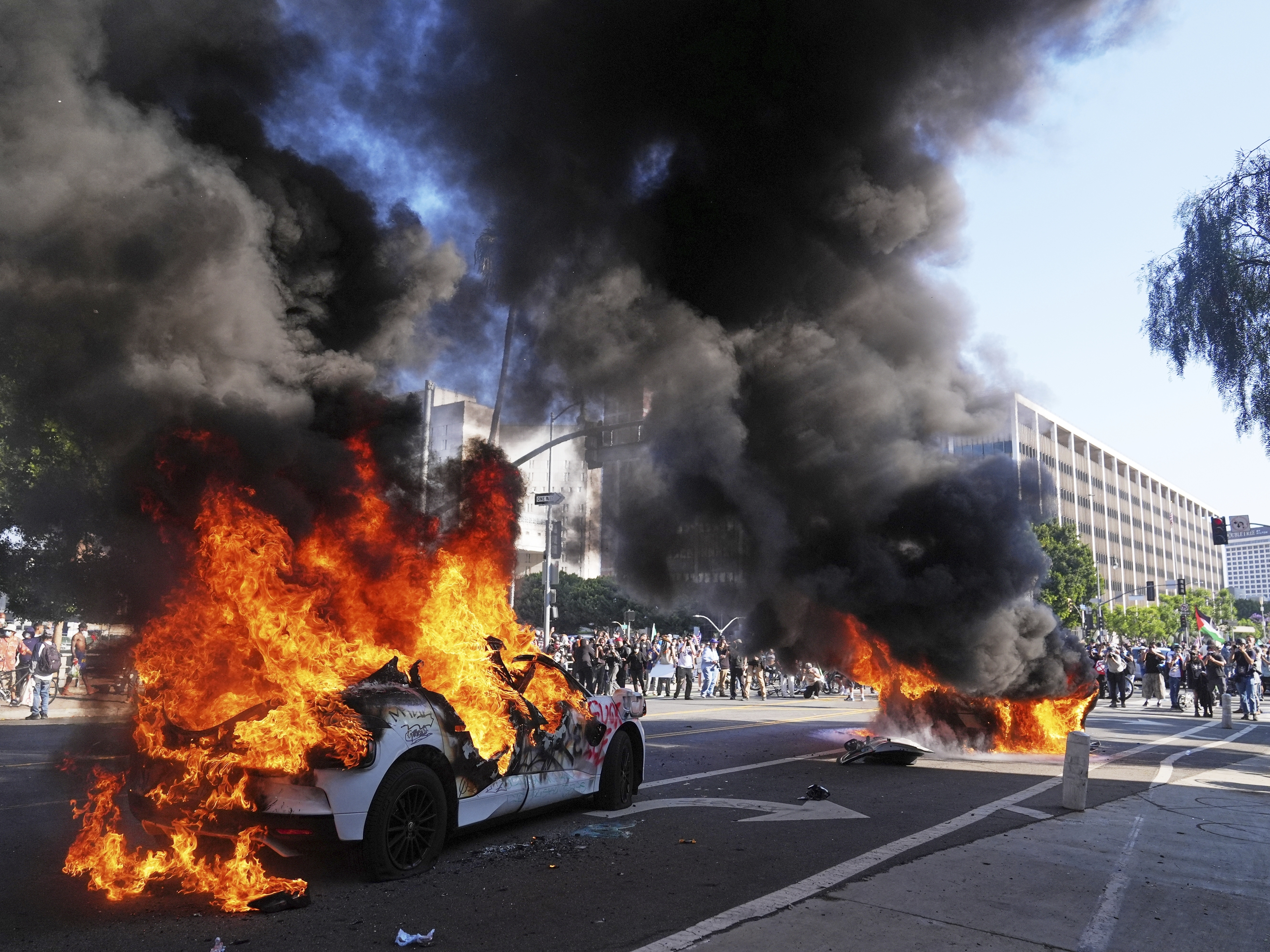 caption: Two Waymo taxis burn near the metropolitan detention center of downtown Los Angeles, Sunday, June 8, 2025, following last night's immigration raid protest.<br>