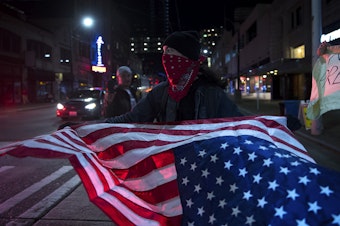 caption: A protester who declined to be named waves an upside down American flag while standing in traffic along with other protesters following the 'Impeach and Remove Rally' on Tuesday, December 17, 2019, in Seattle.
