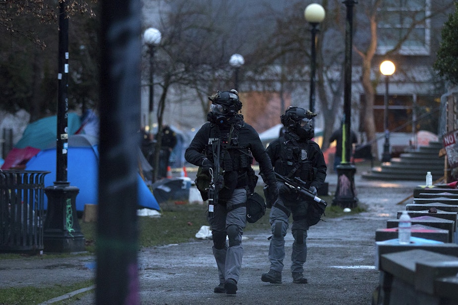 caption: Seattle police officers walk through an encampment of unhoused community members dressed in riot gear shortly after dawn on Friday, December 18, 2020, at Cal Anderson Park in Seattle. 