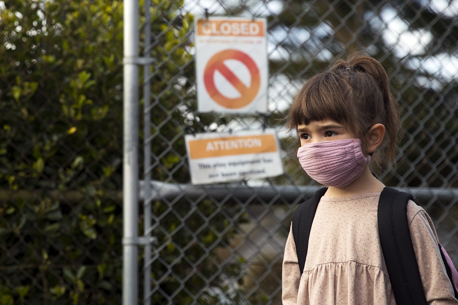 caption: Rooney Walkingstick, a kindergarten student at Northgate Elementary School, waits in line to check into school as flyers relating to Covid-19 closures and sanitation of the playground are visible in the background on a nearby fence, on Monday, April 5, 2021, on the first day of in-person learning at the school in Seattle. "I'm a little scared and a little excited," Rooney said.