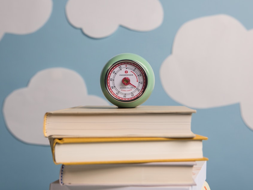 A sea-foam green egg timer set to 20 minutes sits atop a stack of closed books in front of a light blue backdrop covered in cutouts of white clouds.
