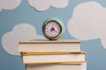 A sea-foam green egg timer set to 20 minutes sits atop a stack of closed books in front of a light blue backdrop covered in cutouts of white clouds.