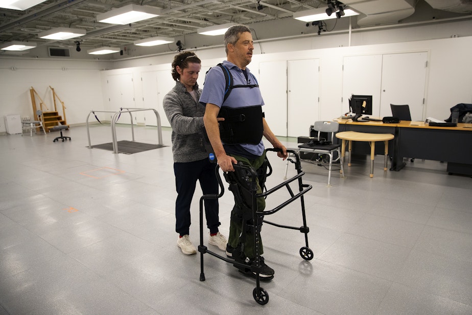 caption: Jon Schlueter, who became injured in a diving accident, walks in an eksobionics device with the help of Rich Henderson, a graduate student and physical therapist, on Monday, April 15, 2024, at Wallace Hall on the University of Washington campus in Seattle. 