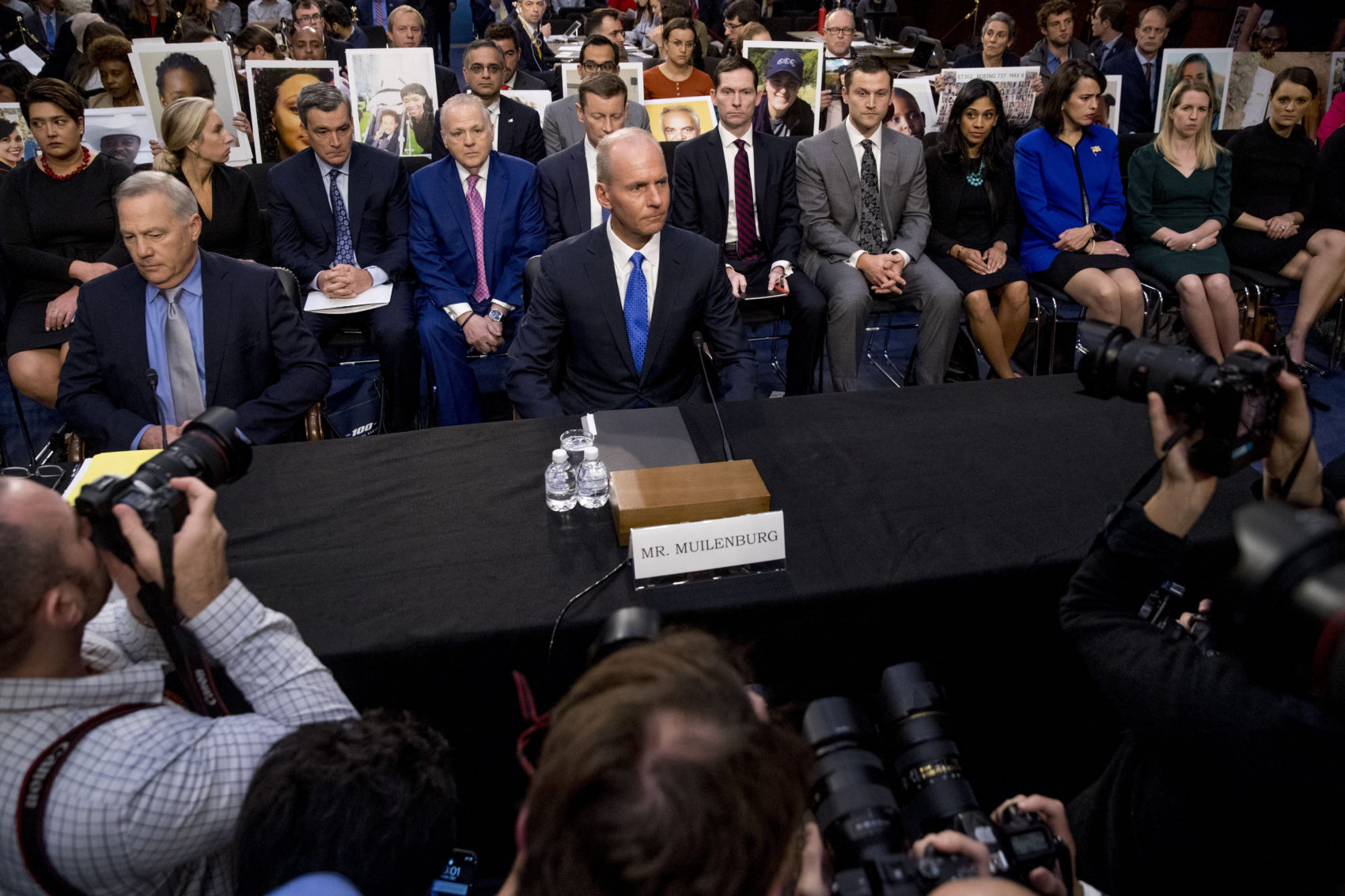 caption: Family members of those killed in the Ethiopian Airlines Flight 302 and Lion Air Flight 610 crashes hold photos of their family members behind Boeing Company President and Chief Executive Officer Dennis Muilenburg, center, and Boeing Commercial Airplanes Vice President and Chief Engineer John Hamilton. (Andrew Harnik/AP)