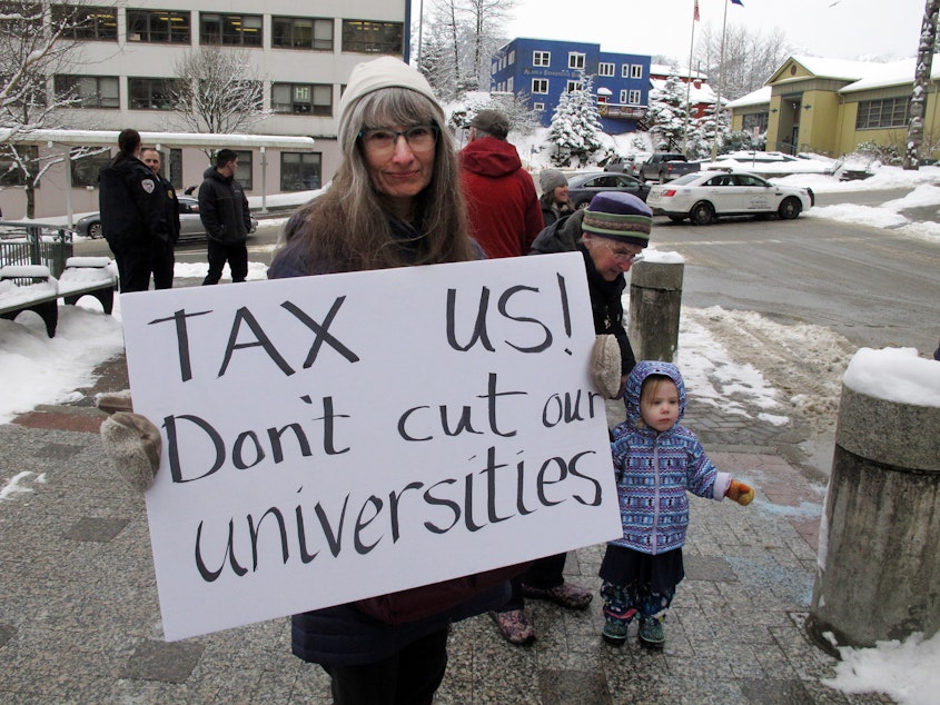 caption: Luann McVey holds a sign before the start of a rally held in support of the Alaska university system on Feb. 13 in Juneau, Alaska.