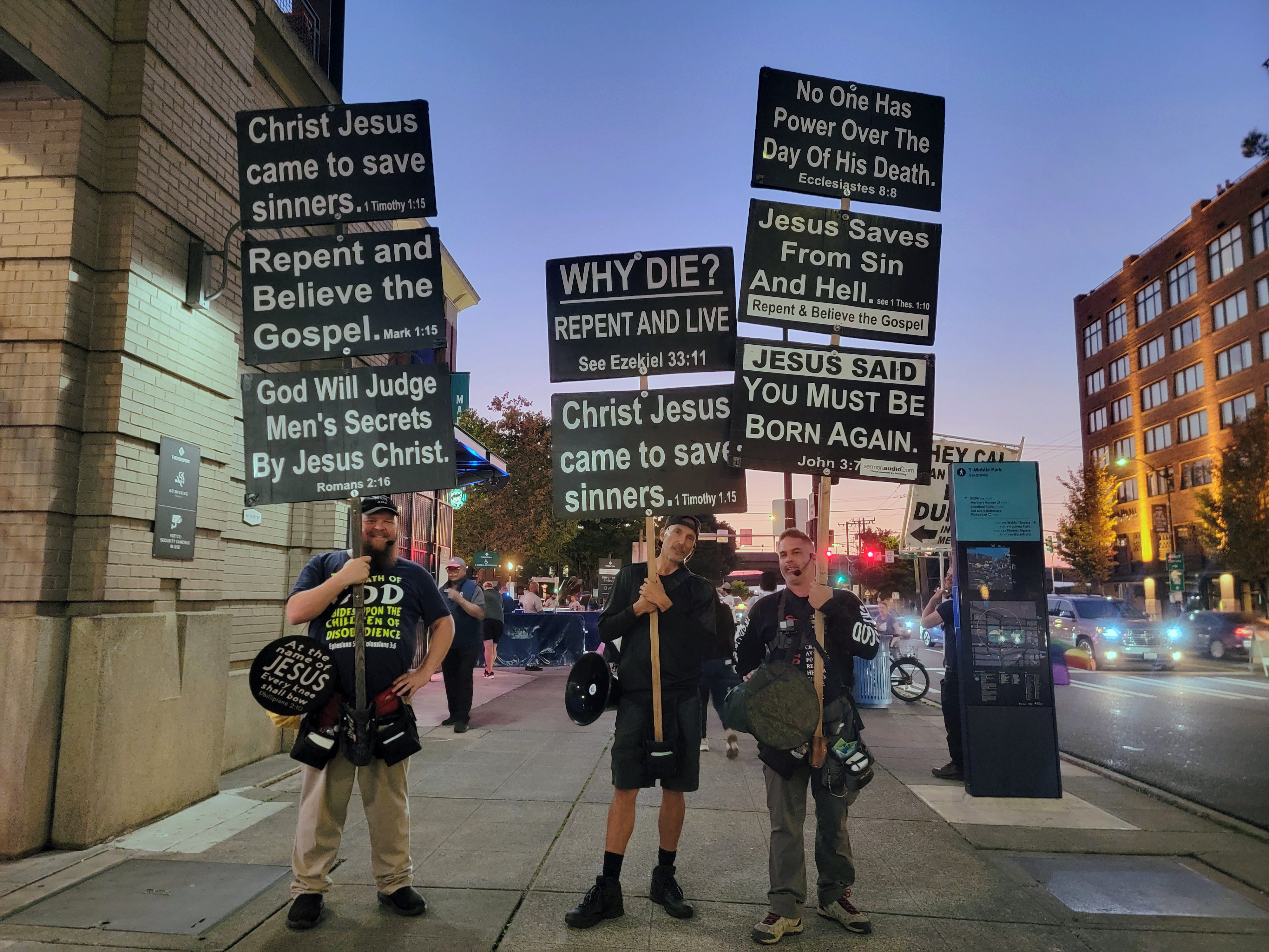 caption: Justin Symons, Thomas C., and Ron Cardiel (right) stand with their signs outside the ballpark on Wednesday, Sept. 24, 2025.