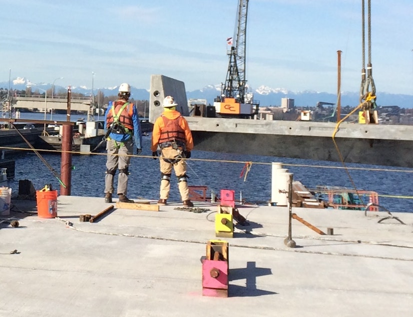 caption: Two construction workers wait to move a deck panel into position. The panel is one of hundreds that will make the road surface of the new 520 bridge. 