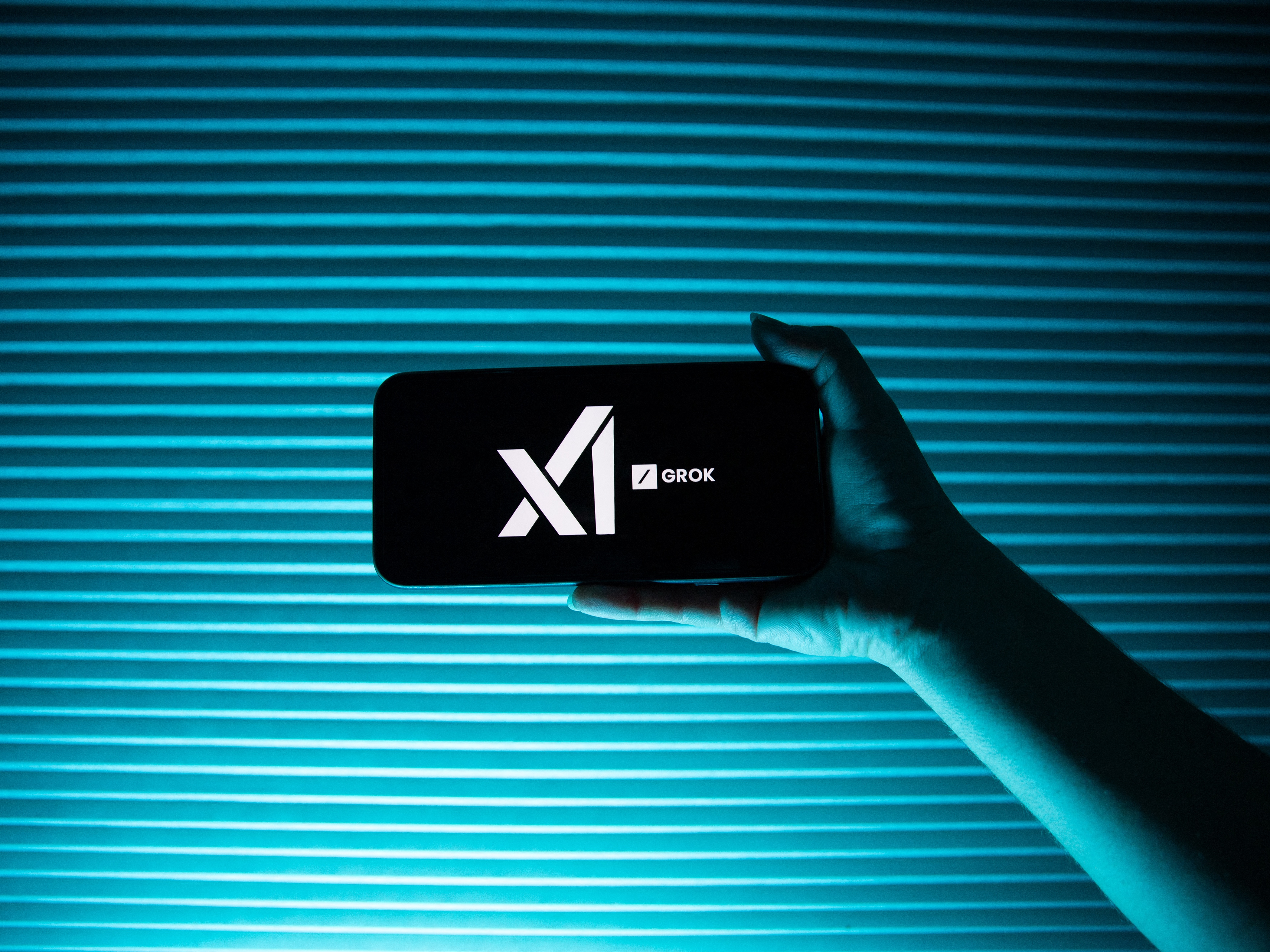 caption: A person holds a telephone displaying the logo of Elon Musk's artificial intelligence company, xAI  and its chatbot, Grok.
