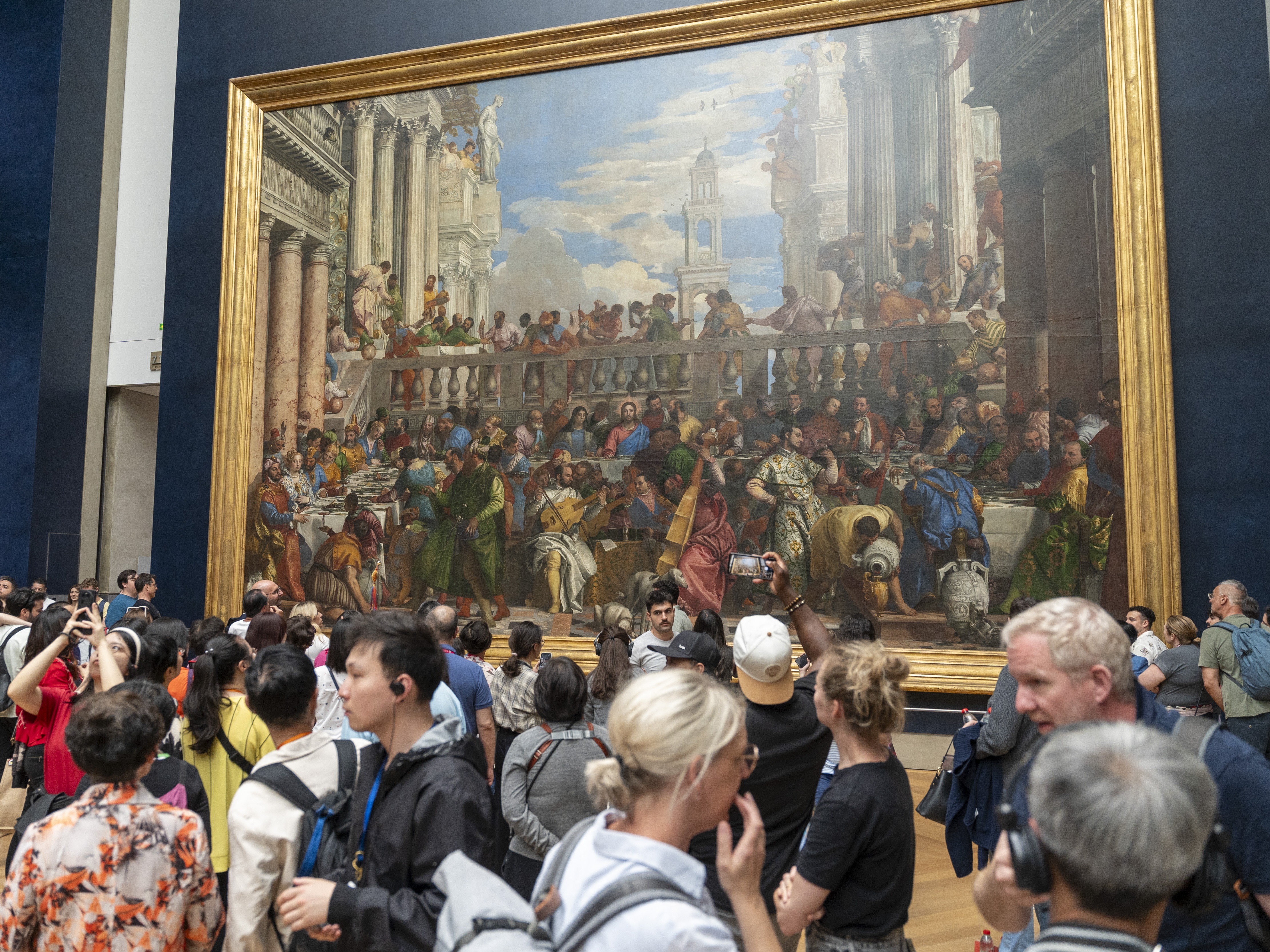caption: Louvre visitors in front of <em>The Wedding Feast at Cana</em> by Paolo Veronese in June 2024. The painting is among several masterworks that may soon cease to play "second fiddle'" to the Mona Lisa.