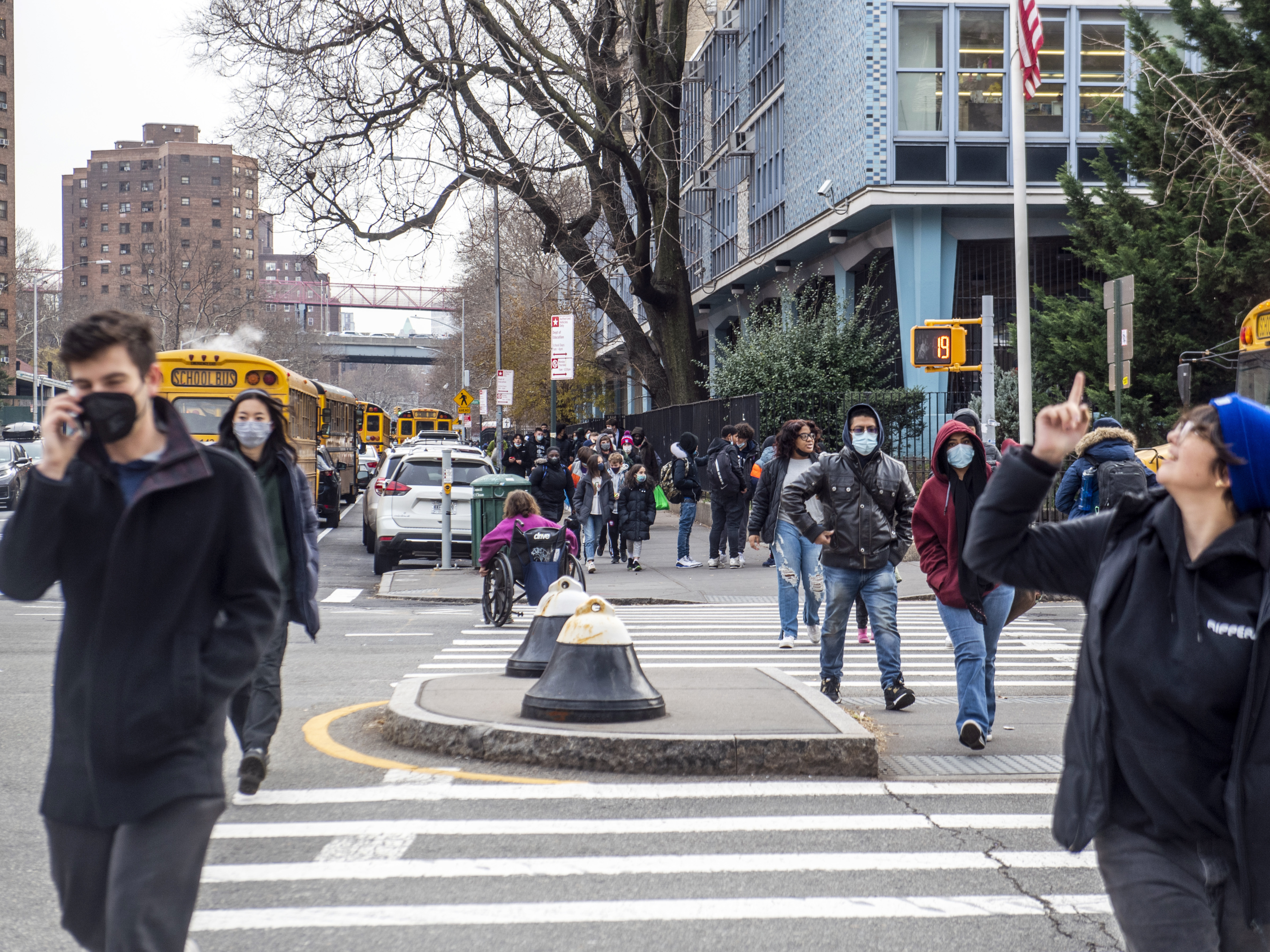 caption: Pedestrians cross Houston Street as students wearing masks leave the New Explorations into Science, Technology and Math (NEST+m) school in the Lower East Side neighborhood of Manhattan on Tuesday, Dec. 21, 2021, in New York.