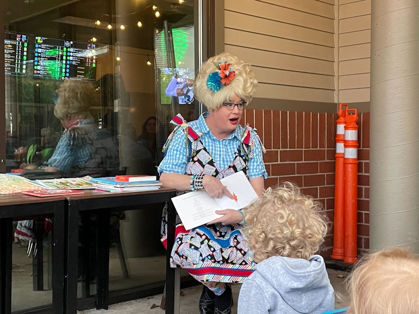 caption: Sylvia O'Stayformore, in a blue checkered shirt and bingo jumpsuit, reads a picture book to children
