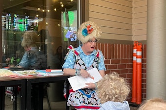caption: Sylvia O'Stayformore, in a blue checkered shirt and bingo jumpsuit, reads a picture book to children