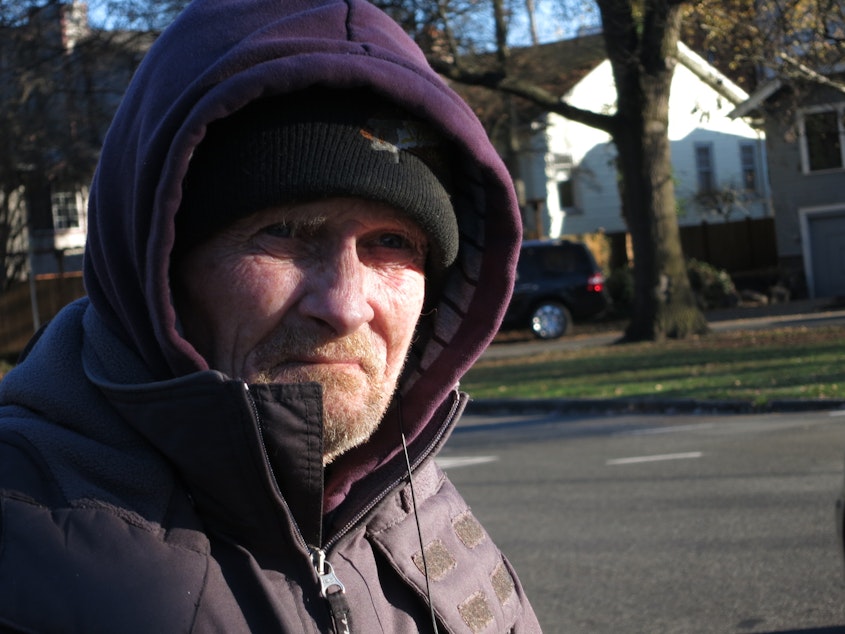 caption: Michael Thompson stands by the NE 65th St. exit from I-5 with a sign asking for help. He suggests providing food, warm clothing, and work to panhandlers. 