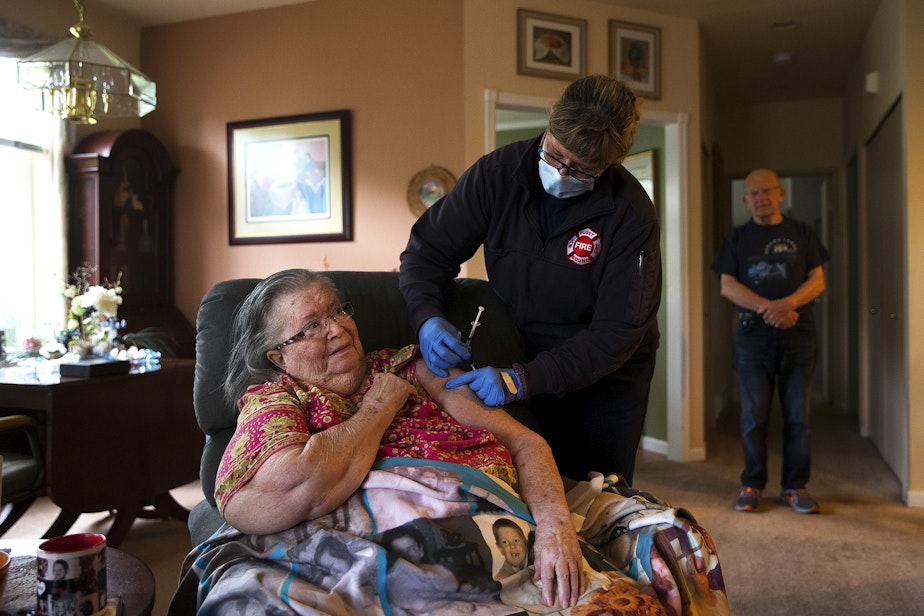 caption: Puget Sound Regional Firefighter Nikki Smith administers a Moderna Covid-19 vaccine for Connie Palmer, as her husband Steve Palmer, right, observes, on Monday, May 24, 2021, at their home in Kent.