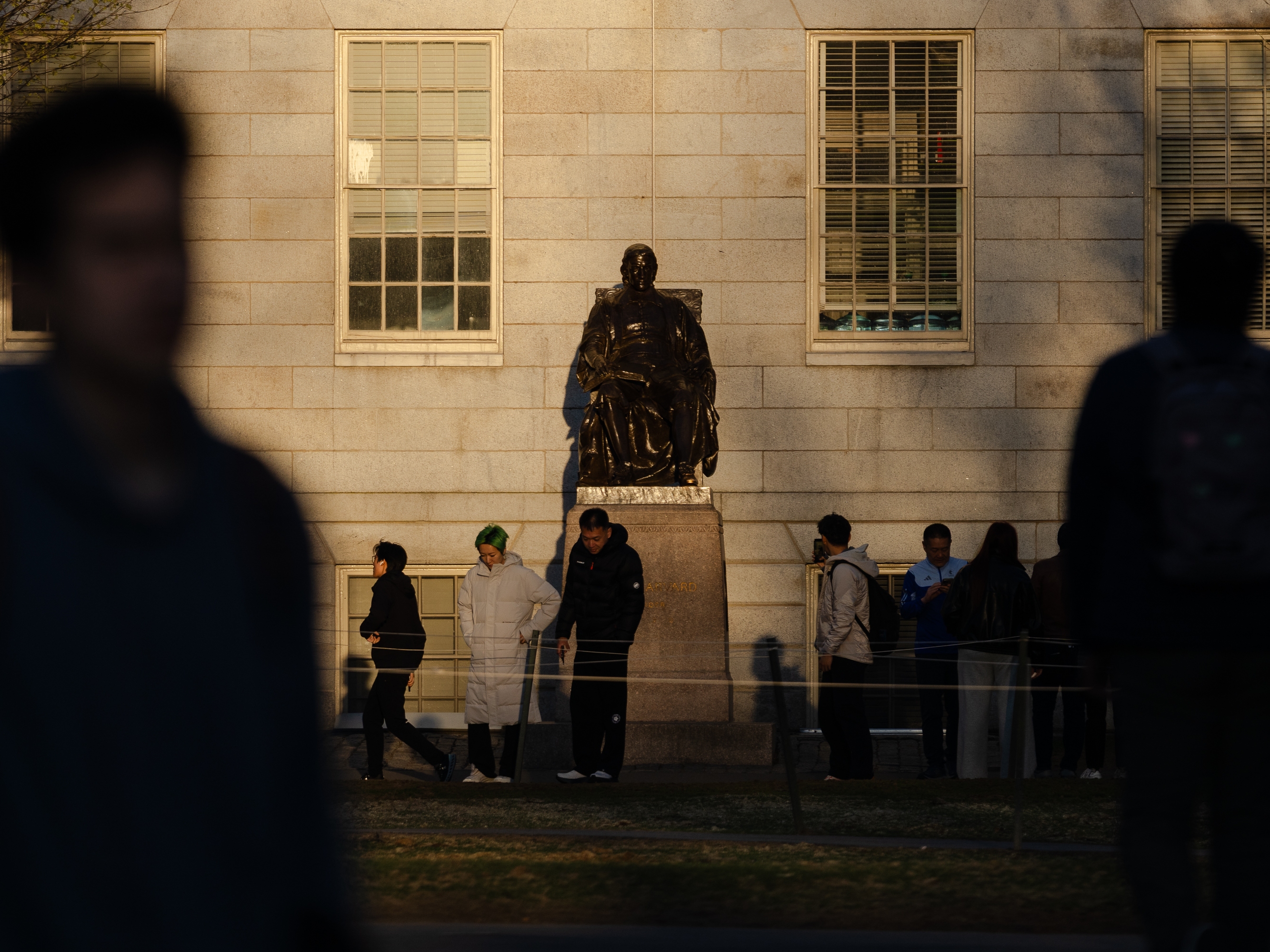 caption: People gather to take photos with the John Harvard statue at Harvard University. President Trump made another threat to Harvard's tax-exempt status in a social media post on Friday.