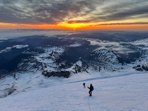 caption: The sun rises over the Central Cascades as the roped-up adventurers ascend to the summit of Mount Rainier in late May.