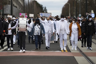 caption: James Bible, the Ellis family's attorney, center, marches silently along with about 200 others to the beat of a single drum, played by drummer Elyshau Wilson, left, in honor of 33-year-old Manuel Ellis who was killed by Tacoma police one year ago, on Sunday, February 28, 2021, along Martin Luther King Jr. Way in Tacoma.