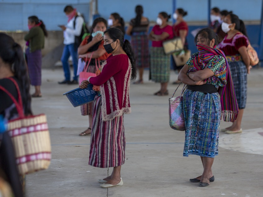 caption: Women wearing face masks stand at a safe distance to help curb the spread of the new coronavirus, as they wait for food assigned to their children outside a school in Xesuj, Guatemala, where many residents depend on remittances, largely from the U.S. The fallout from the pandemic is cutting into the financial lifelines for people across Latin America, Africa and Asia.