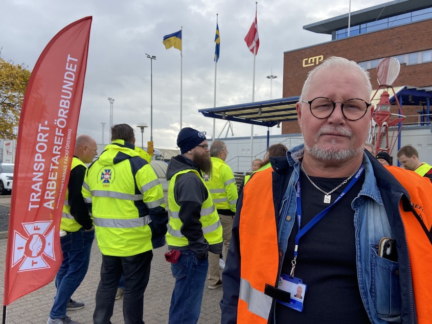 caption: Goran Larsson, a cargo ship inspector, poses next to the Transport Workers' Union flag at the Malmo port on Nov. 7. Dockworkers are refusing to load or unload Teslas at this port and all others across the country.