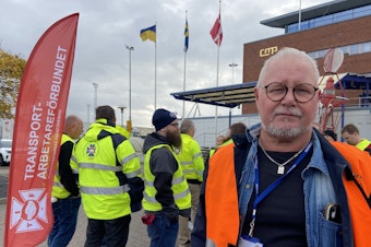 caption: Goran Larsson, a cargo ship inspector, poses next to the Transport Workers' Union flag at the Malmo port on Nov. 7. Dockworkers are refusing to load or unload Teslas at this port and all others across the country.