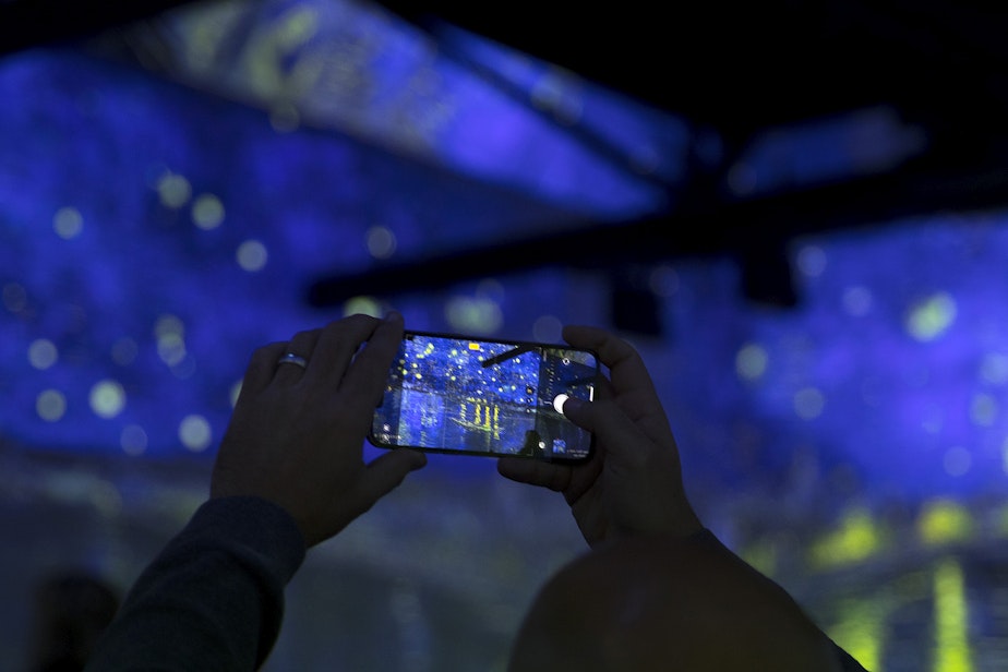 caption: A person takes a photograph with their cellphone as various projections of Van Gogh's work are displayed in the 360-degree 8,000 square foot immersive room on Wednesday, October 27, 2021, at the exhibit in Seattle's SODO neighborhood.