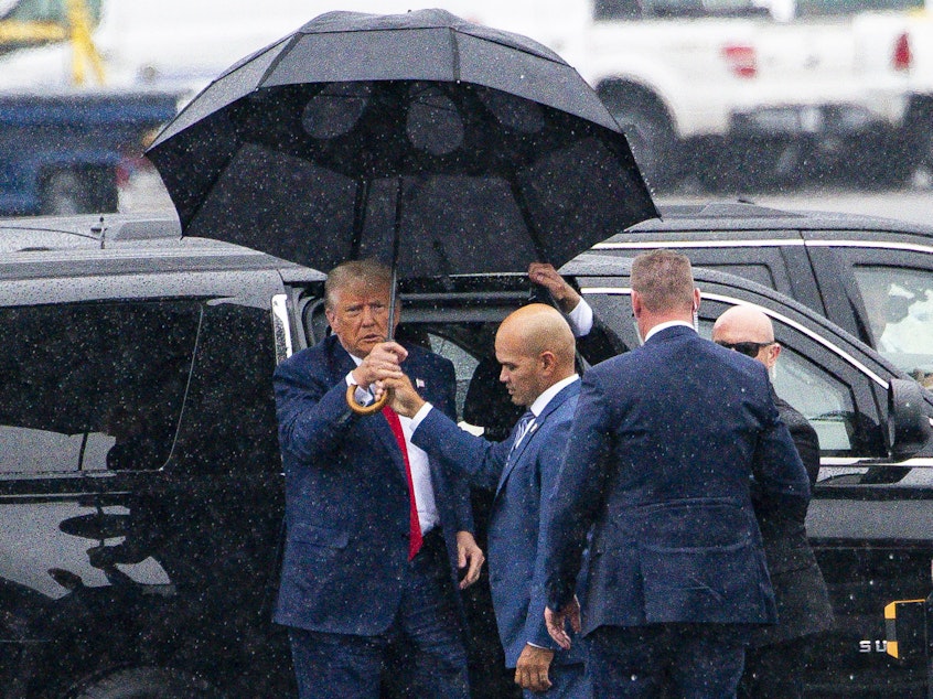 caption: Donald Trump, left, is handed an umbrella from Walt Nauta, his personal aide and co-defendant in a felony case in Florida, on Thursday, the day the former president pleaded not guilty to four felony charges in Washington, D.C.