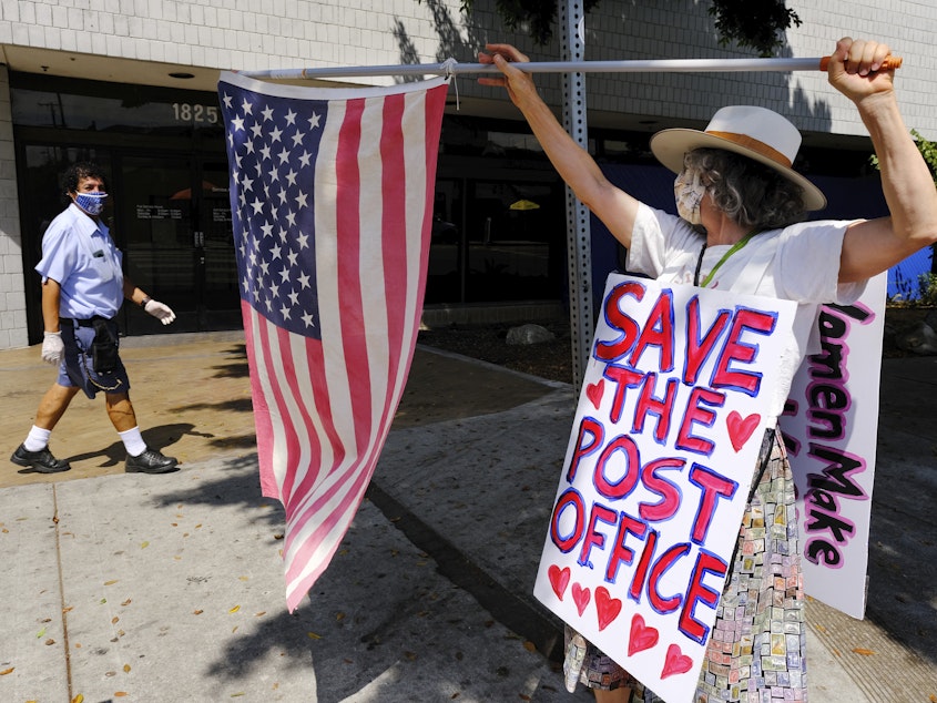 caption: Erica Koesler of Los Angeles demonstrates outside a USPS post office as a postal worker walks by in the background on Saturday. The USPS has warned states coast to coast that it cannot guarantee all ballots cast by mail for the November election will arrive in time to be counted, even if mailed by state deadlines.