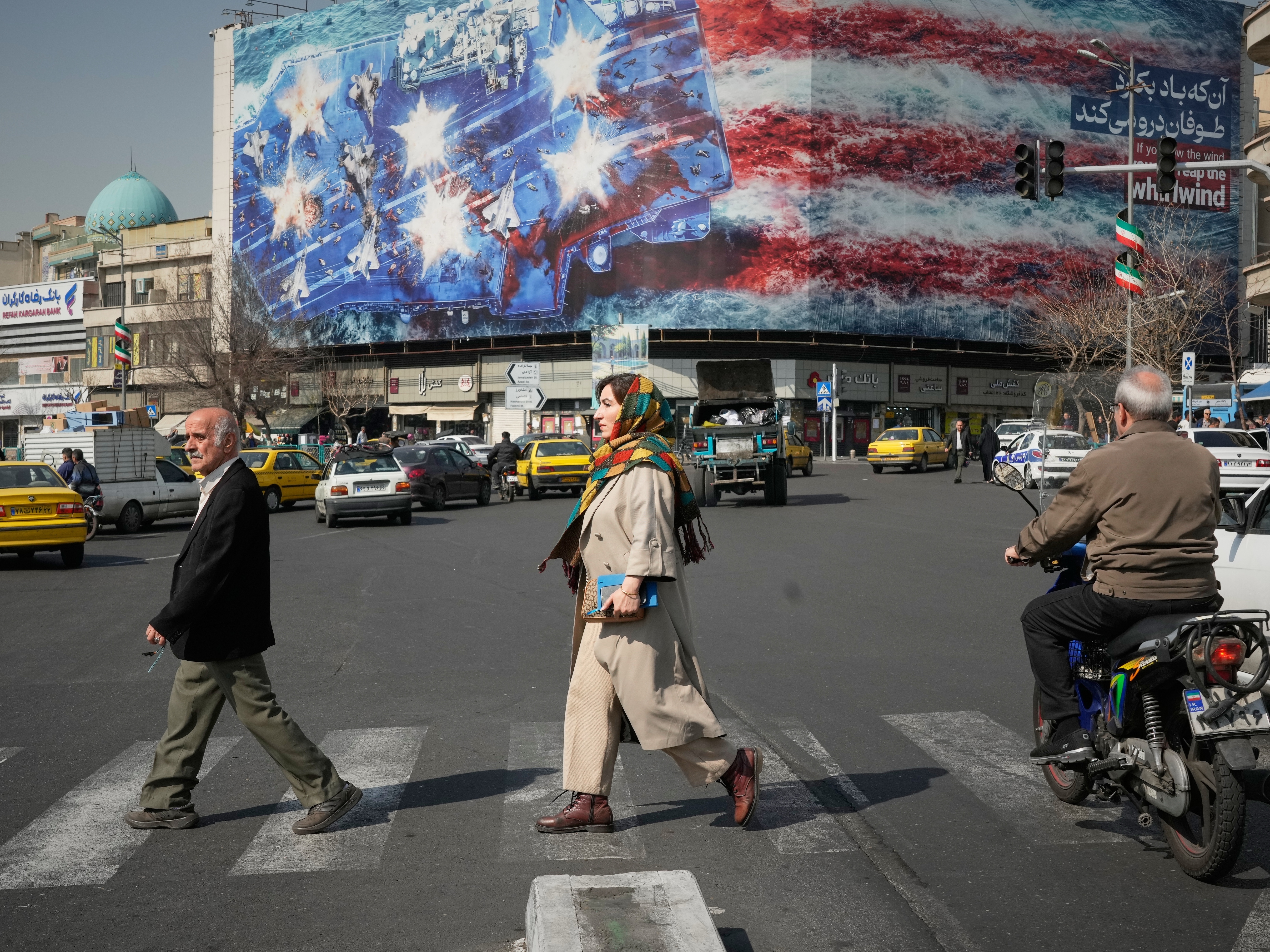 caption: Pedestrians walk past a billboard depicting a U.S. aircraft carrier with damaged fighter jets on its deck and a sign in Farsi and English reading, "If you sow the wind, you'll reap the whirlwind," at Enqelab-e-Eslami (Islamic Revolution) Square in Tehran, Iran, Sunday, Feb. 22, 2026.