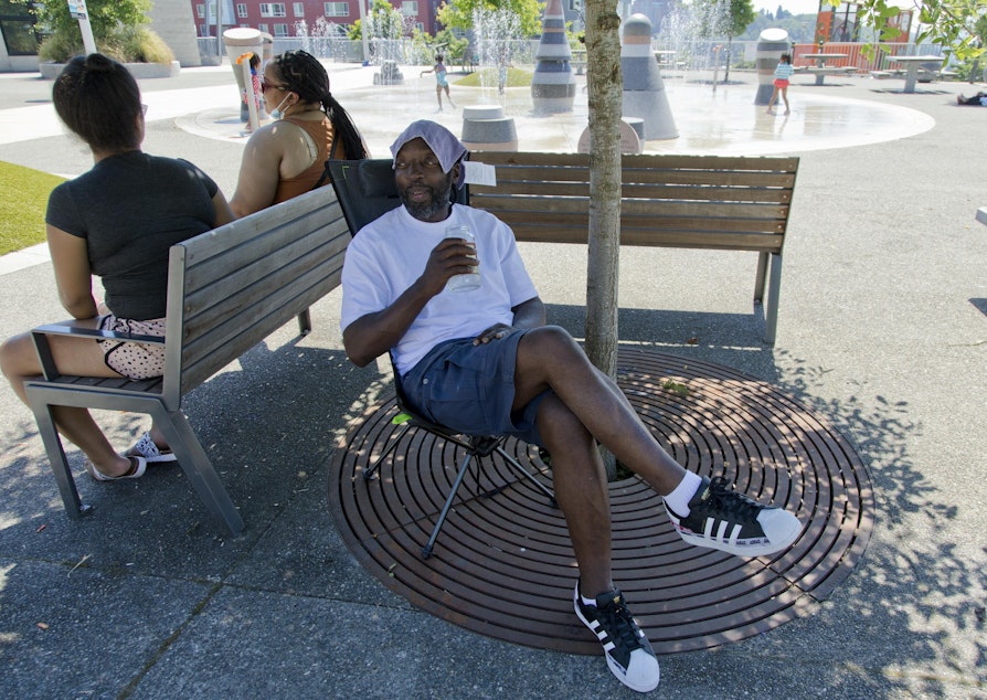 caption: Melvin O'Brien waits in the shade in Yesler Terrace Park while his children play in the spray park during a heat wave hitting the Pacific Northwest, Sunday, June 27, 2021, in Seattle. 